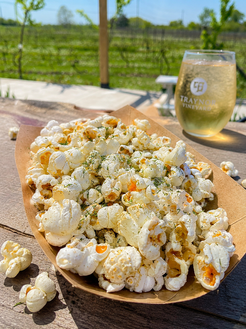 Garden Herb Popcorn at Traynor Family Vineyard - popcorn tossed with dried dill, oregano, garlic, and flaky salt in a kraft tray, with a glass of Traynor sparkling wine and the Hillier vineyard behind