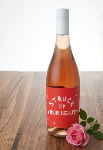 Wine bottle with a red label reading 'Struck by Your Beauty' on a wooden surface with a pink rose.