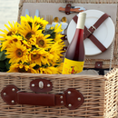 "A wicker picnic basket with leather straps, containing a bouquet of bright yellow sunflowers, a bottle of Inclusion Orange wine with its yellow label featuring a whimsical red dog riding a bicycle, and picnic essentials such as white plates, cutlery, and a corkscrew neatly arranged inside."