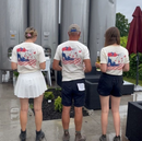 "Three individuals standing with their backs to the camera in front of large stainless steel wine tanks. They are wearing matching white t-shirts featuring a colorful design of a blue figure pouring wine from a red teapot into cups, with a red and white checkered pattern. The person in the middle wears dark shorts and a cap, while the individuals on either side wear skirts and shorts, respectively. One of the people on the right is holding a bottle of wine. The setting appears to be an outdoor winery or pro