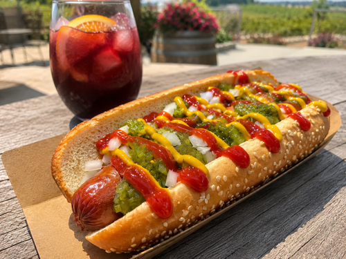 The Classic Dog at Traynor Family Vineyard - an all-beef hot dog on a sesame bun with mustard, ketchup, sweet relish, and onion, with a glass of Traynor red over ice and the Hillier vineyard behind