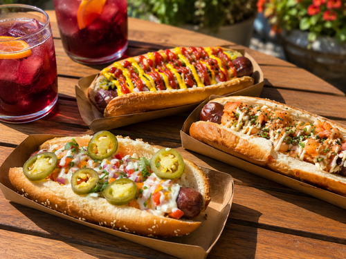 Three Traynor gourmet hot dogs - Classic, Mango Salsa, and Kimchi - with red wine coolers on the patio table at Traynor Family Vineyard, Hillier, Prince Edward County